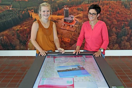 Luisa Möser und Stefanie Heimes (von links) von der Tourist-Information Lennestadt-Kirchhundem in der Zweigstelle in Saalhausen. Im Hintergrund eine Luftaufnahme des Ausflugslokals und Aussichtsturms "Hohe Bracht" auf einer 587,9 Meter über Normalnull befindlichen Bergkuppe. FOTO: ANDREAS DUNKER