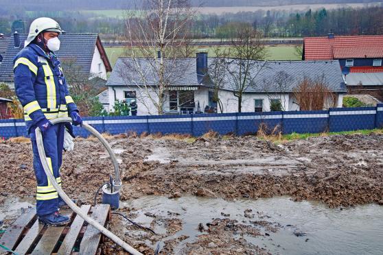 Feuerwehrmann Alveenus Anthonipillai (genannt: „Alwin“) mit einer Saugpumpe beim Absaugen des Oberflächenwassers am Rande des Neubaugebiets "Osterdorf" in Echthausen. – Im Hintergrund ist die Terrasse des Wohnhauses der Familie Münstermann mit weiteren Feuerwehrleuten zu sehen. FOTO: ANDREAS DUNKER