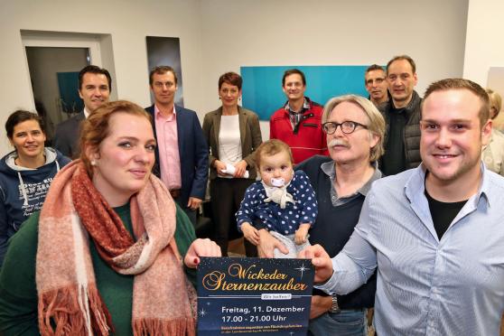 Die Malteser-Mitarbeiter Clara Hüttenbrink (links) und Julian Demiet (rechts) präsentierten zusammen mit WIR-Sprecher Dr. Clemens Frigge (Mitte) – hier mit Enkelkind – ein neu entworfenes Plakat zu der Aktion „Wickeder Sternenzauber – Wir helfen“. Die Hilfsaktion der Händler ist zugunsten bedürftiger Flüchtlinge in der „Zentralen Unterbringungseinrichtung“ (ZUE) in Wimbern. Im Hintergrund (von links):  Gabriela Kimpel, Volksbank-Direktor Holger Georg Schwarz, Rechtsanwalt Sascha Hesse, Apothekerin Martina Brennecke, Dirk Reinke, Rüdiger Papenberg, Detlef Lojewski und Margret Schulte (verdeckt). FOTO: ANDREAS DUNKER